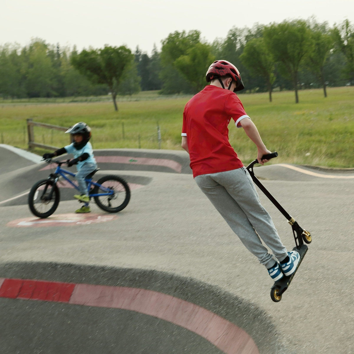 Junge springt mit dem Stuntscooter, dahinter fährt ein Kind Fahrrad